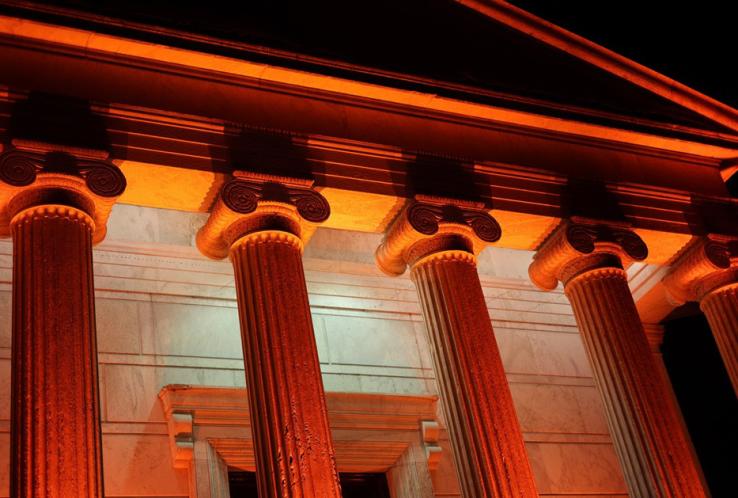The tops of five Ionic columns on a building are lit up with orange lights.