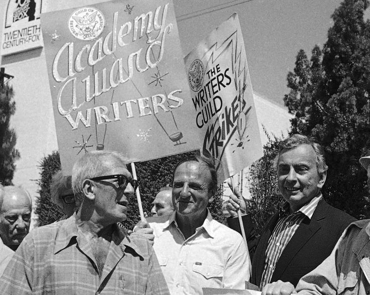Black-and-white photo of three men in a crowd holding signs reading "Academy Award writers" and "The Writers Guild Strikes."