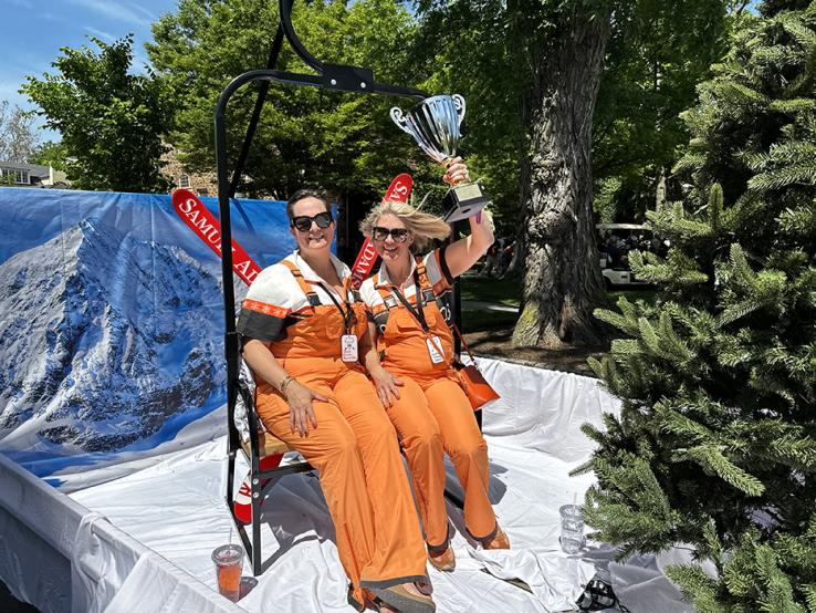 Two women wearing orange overalls; one is holding a trophy over her head.