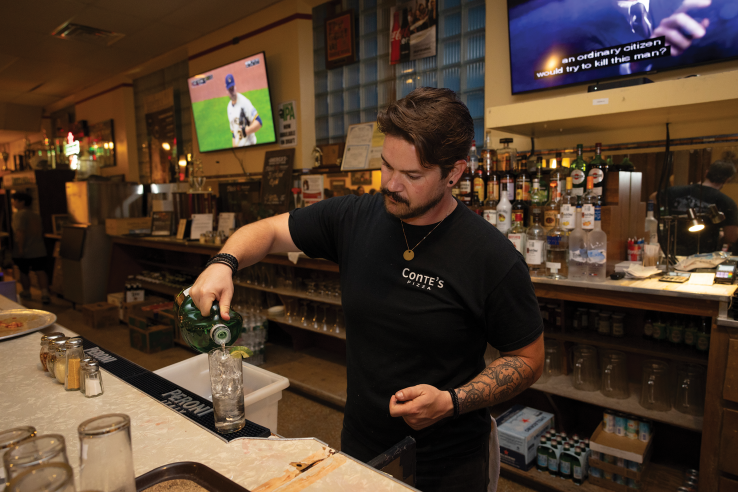 A bartender pours liquor into a glass full of ice behind Conte's bar.