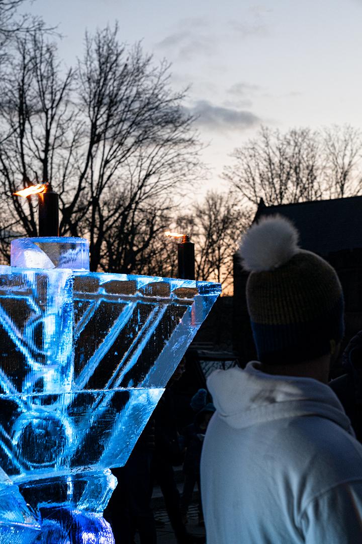 A view of the ice menorah showing it's about as tall as the person standing beside it in a winter hat.
