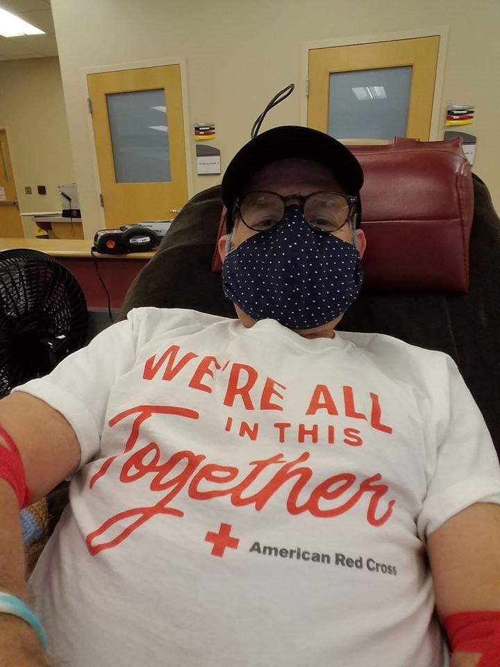 Donating blood, Doug Schutte ’75 wears a cloth mask and a T-shirt that reads, "We're all in this together."