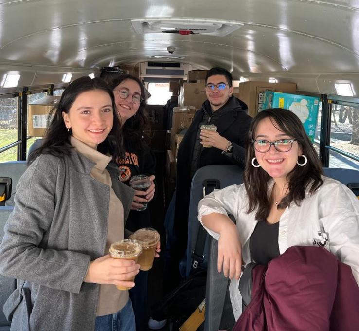 Four students hold cups of coffee on the bus to New York, with boxes stacked behind them.