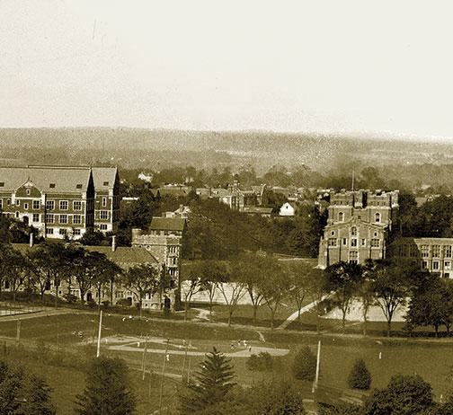 Large science labs Palmer and Guyot are pictured with the rooftops of Prospect Avenue clubs between them. A ballgame is underway at Brokaw Field, today the site of Whitman College. Stately trees on Elm Drive screen Patton dormitory, with tennis courts on 