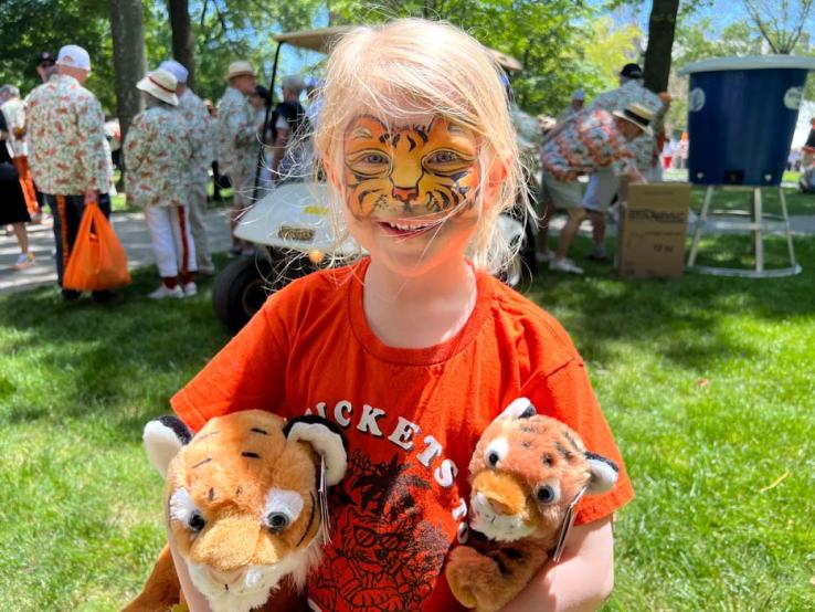 A six-year-old girl with tiger faceprint, an orange shirt, and a plush tiger under each arm.