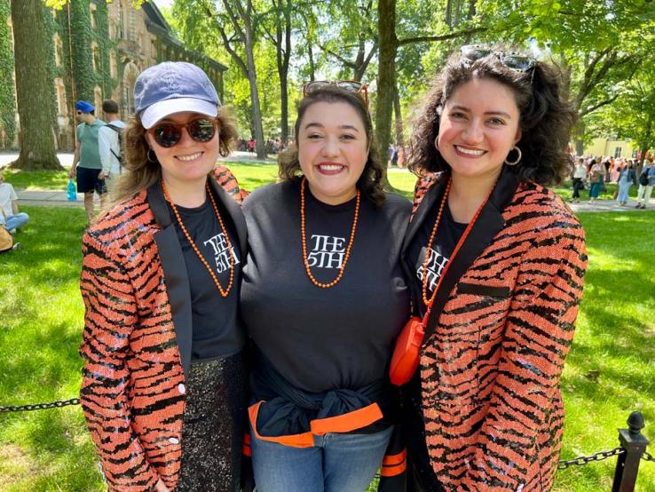 Three women wearing orange martini gras beads and T-shirts reading "The 5th"; the two on the outside are wearing sequined, tiger-striped jackets.