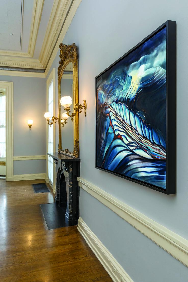 This is a photo "Pools," an abstract painting with lots of blue, hanging in Prospect House on a gray wall with a chair rail and crown moulding, next to a fireplace with a gilded mirror over it.