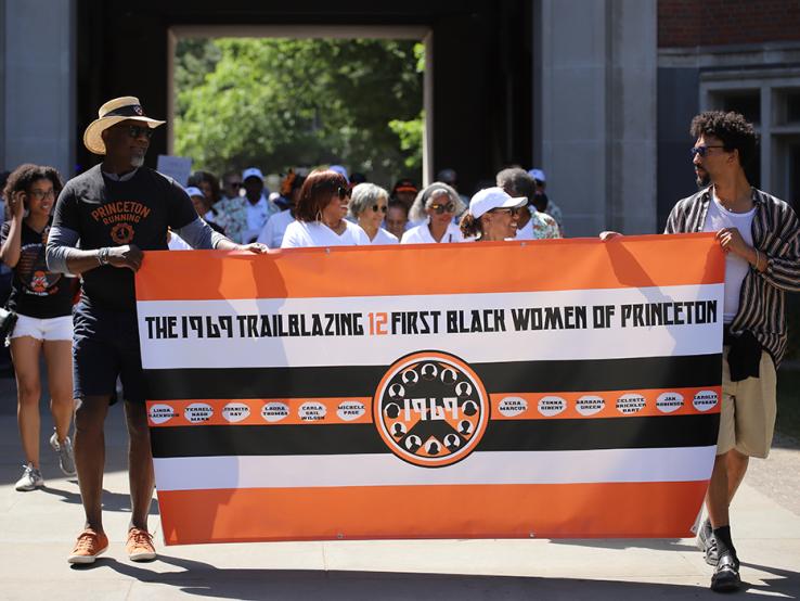 Two guys carry a banner reading "The 1969 Trailblazing 12 First Black Women of Princeton."