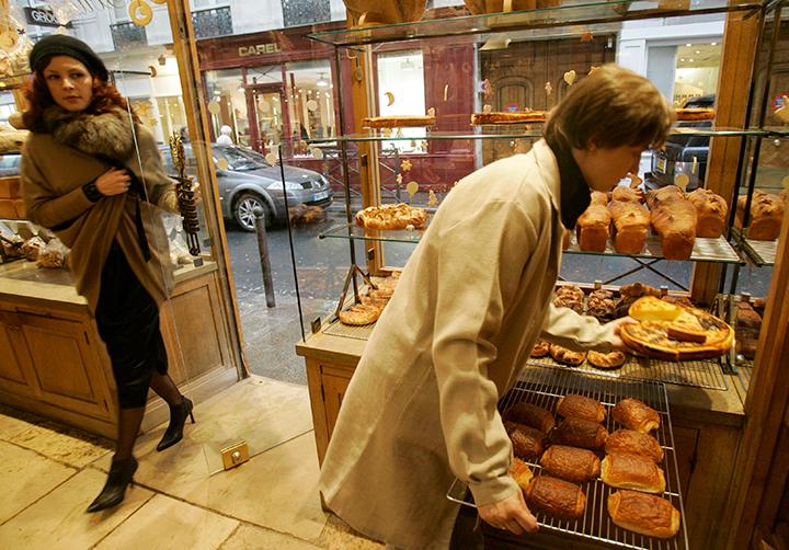 A woman walks into a bakery where another woman leans over a tray of pastries.