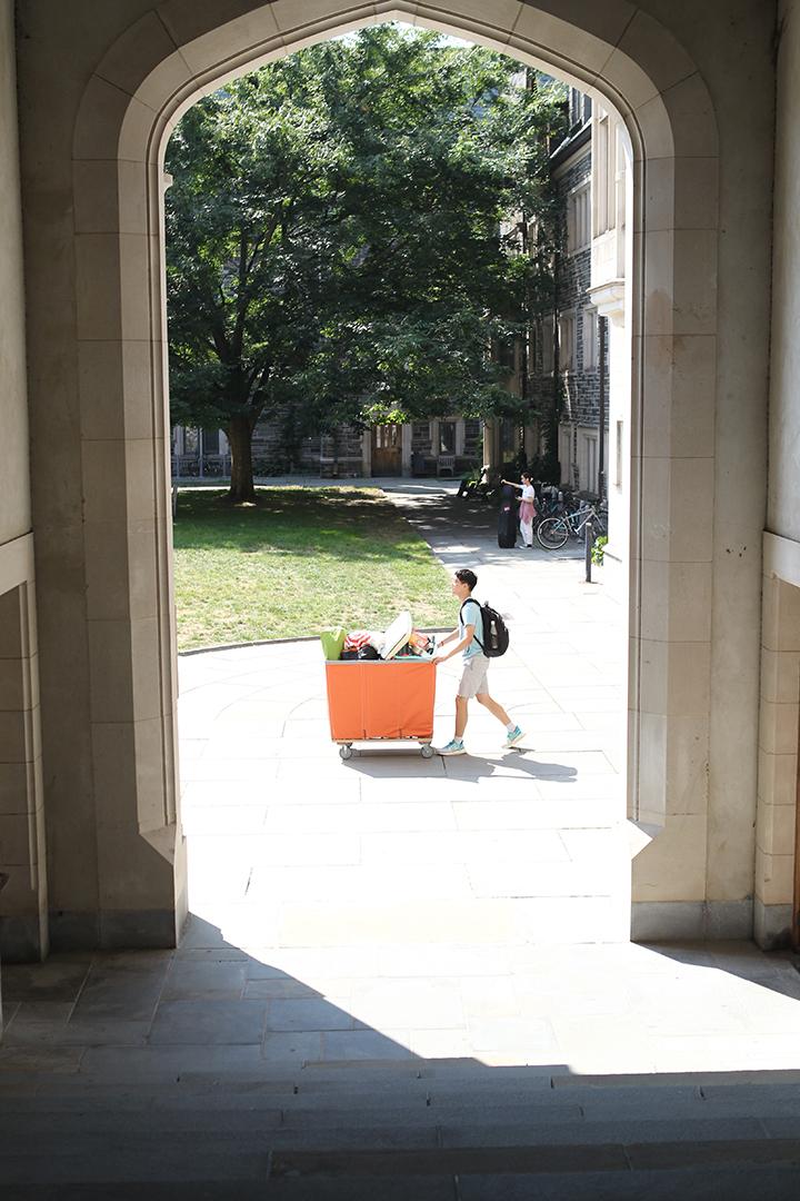 A student pushes a large orange bin on wheels past a stone archway.