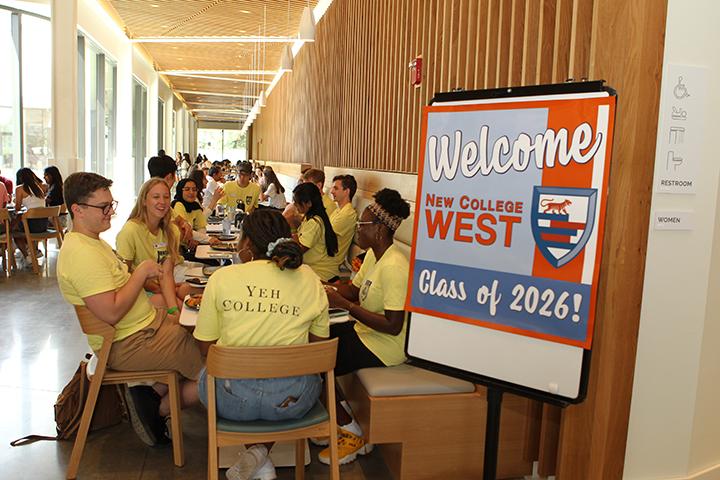 Students in yellow shirts sit around tables next to a sign reading, "Welcome Class of 2026! New College West."