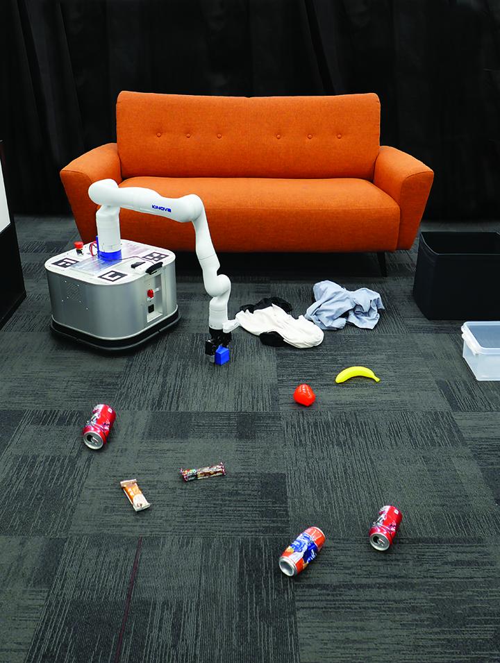 A square robot with a long white arm picks up objects on a carpeted floor in front of an orange couch.