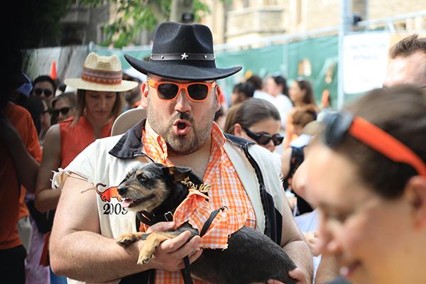 A man in the P-rade wears orange sunglasses and a black cowboy hat and carries a dog.