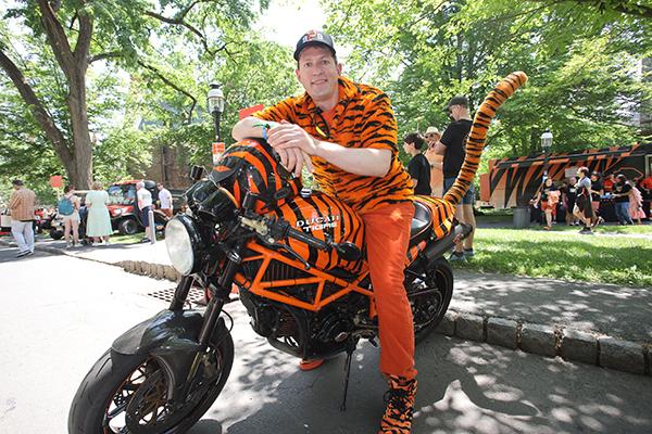 A man in a tiger striped shirt and orange pants leans on a motorcycle covered with tiger stripes that has a long fuzzy tiger tail.