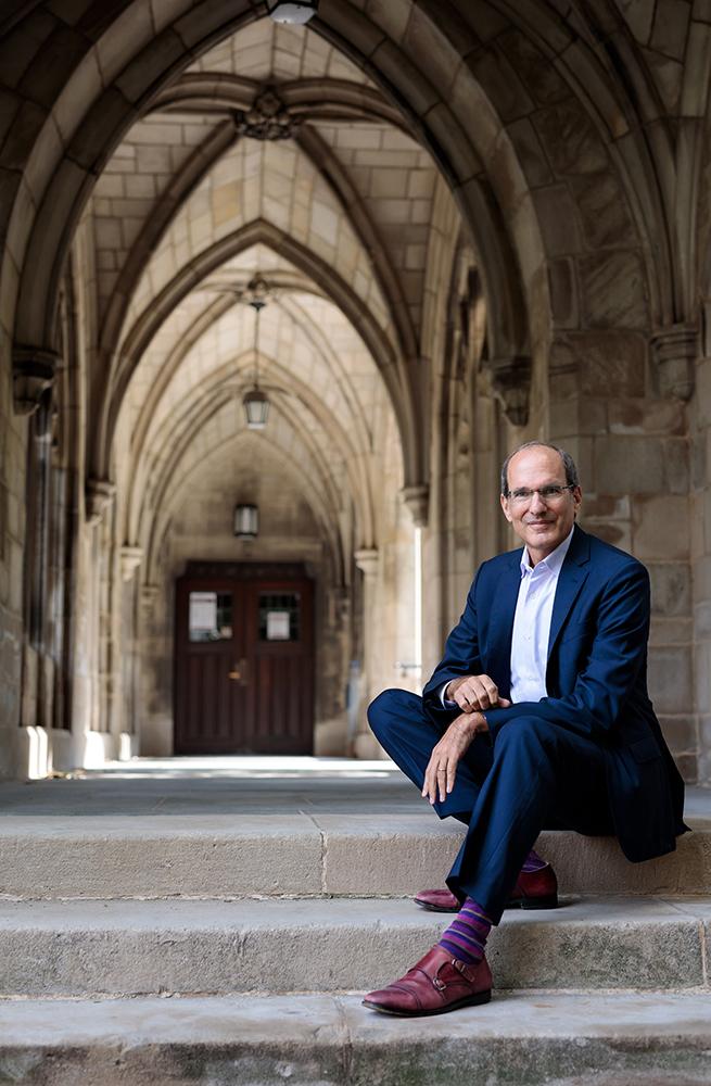 This is a photo of David Nirenberg sitting on the steps of a building with Gothic arches in the background.