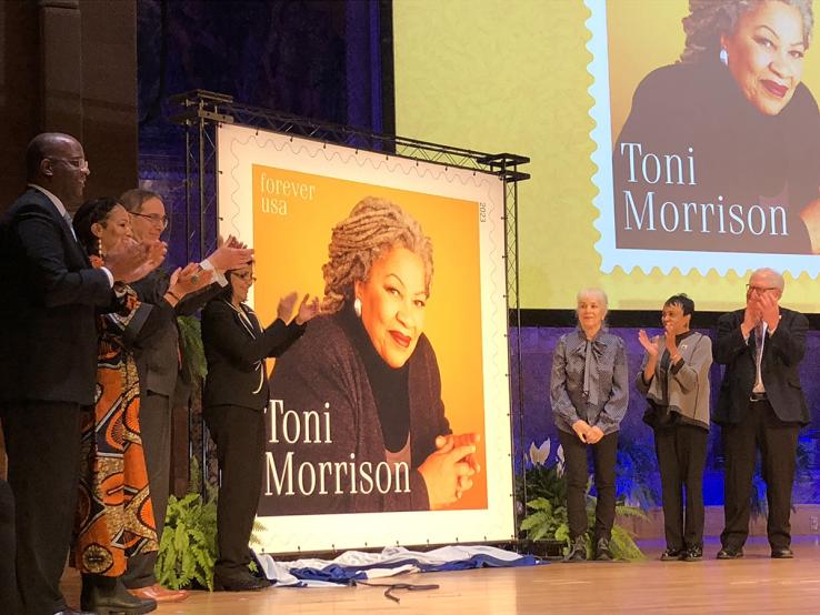 Seven people stand next to a large poster of a postage stamp featuring Toni Morrison