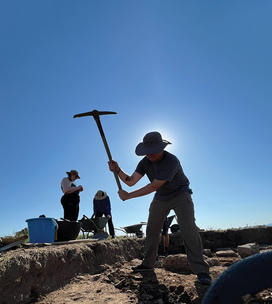 A man swings a pick on an archaeological site, the sun behind his head.