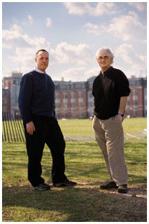 Two men stand on grass with a building far in the background.