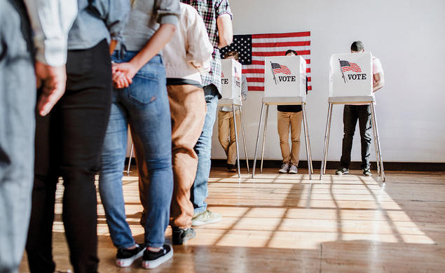 A line of people waiting to vote is shown from behind, from the waist down. People vote in the background.
