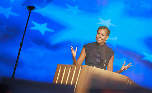 Michelle Obama ’85 speaks at a podium in front of a blue starred background; a teleprompter is in the foreground.