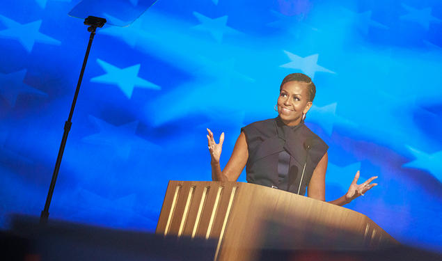 Michelle Obama ’85 speaks at a podium in front of a blue starred background; a teleprompter is in the foreground.