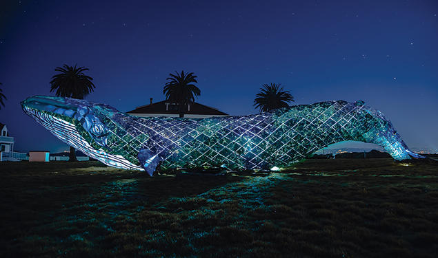 A huge plastic whale, photographed at night, obscures a small building and three palm trees sticking up from the background.
