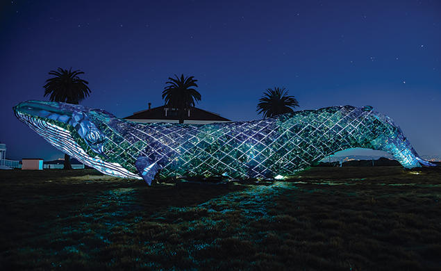 A huge plastic whale, photographed at night, obscures a small building and three palm trees sticking up from the background.