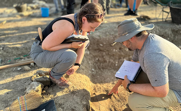 Two people at an archaeological dig examine something in the dirt.