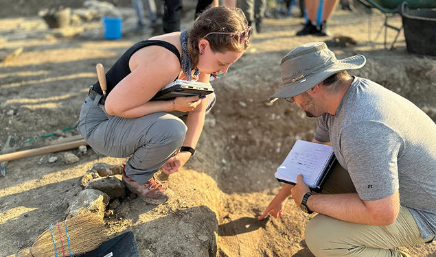 Two people at an archaeological dig examine something in the dirt.