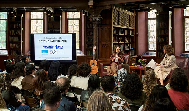 Two women and a guitar next to a screen, speaking to a crowd.