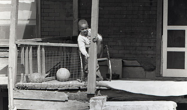 In this black-and-white photo from the 1960s, a boy stands on the porch of a home in rough shape, talking to another boy on the ground.