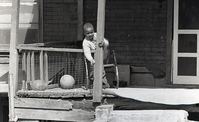 In this black-and-white photo from the 1960s, a boy stands on the porch of a home in rough shape, talking to another boy on the ground.