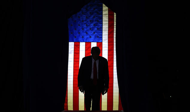 Trump standing in front of a lit U.S. flag surrounded by darkeness