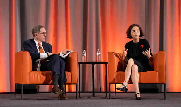 Two people sit on orange chairs on a stage.