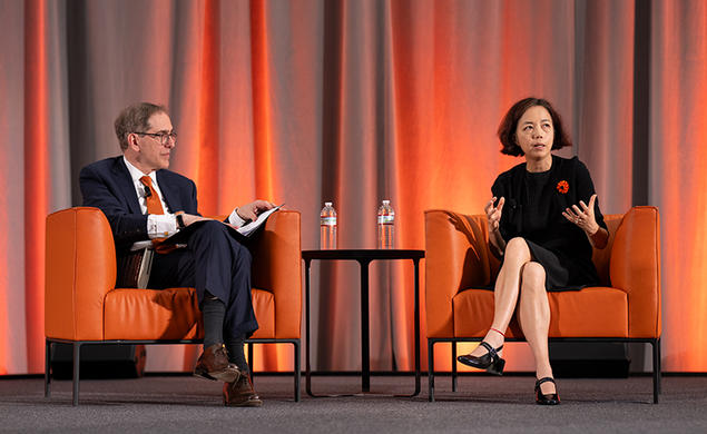 Two people sit on orange chairs on a stage.
