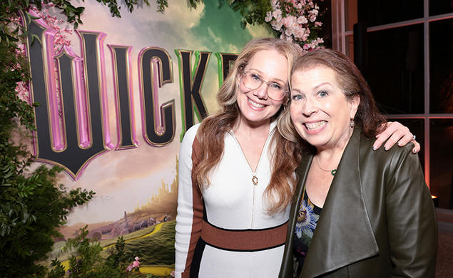 Two women smile for the camera in front of a poster for "Wicked" the movie.
