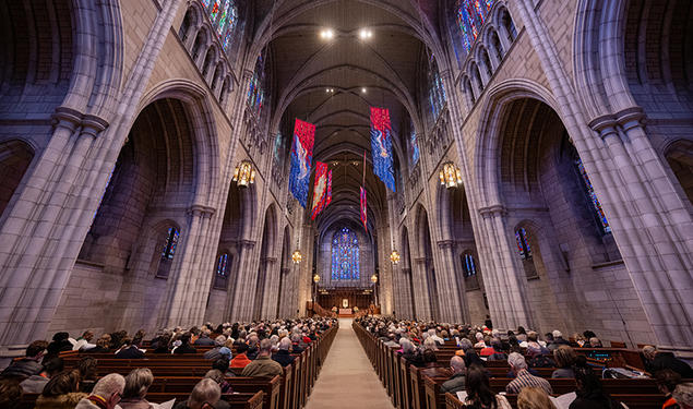 View down the aisle of the Gothic University Chapel, flanked by stone arches and full of people in the pews.