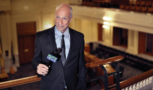 Michael Aron *70 holds a microphone while speaking from an upper level of the NJ statehouse.