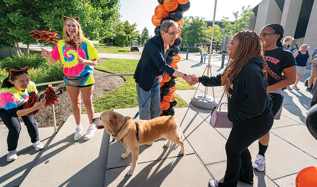 President Eisgruber shakes a woman's hand in front of an orange-and-black balloon arch; his other hand holds the leash of a yellow labrador.