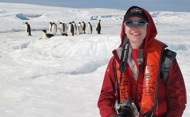 A woman in a red jacket and sunglasses holds a camera in an ice-covered place, while about a dozen emperor penguins stand in the background.