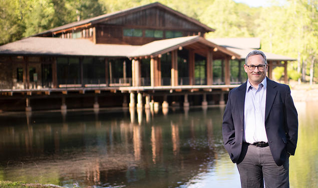 Jason Posnock ’94 stands in front of a pond and a rustic-looking building at the Brevard Music Center.