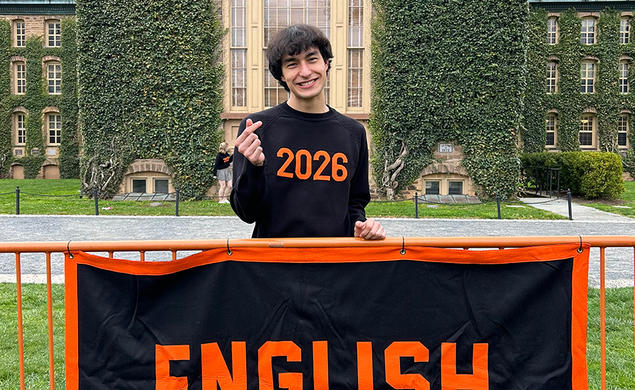 Lauren Blackburn ’26 stands smiling behind a fence with a banner that reads ENGLISH in orange on a black background.