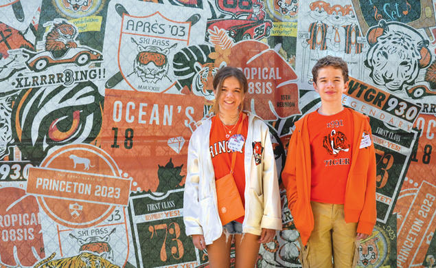Violet and Everett Dutton standing in front of a collage of Class logos at Reunions 2023