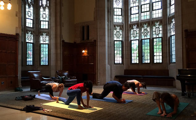 Five people practice yoga in a dim room with Gothic windows.