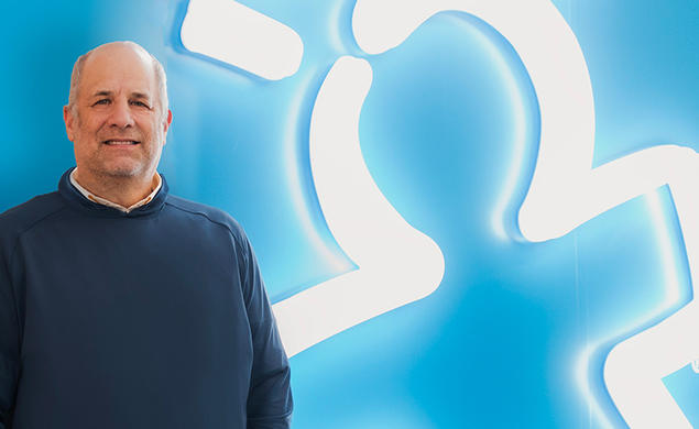 Rick Woldenberg ’81 against a blue background where his company's logo — a person with lines like a lighbulb overhead — is glowing in white neon bulbs.