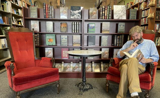 John Kuhner sits at one of two red chairs and reads a book in front of a bookcase in his bookstore.