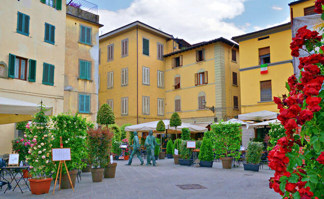 Yellow and pale white buildings surround a plaza with small trees and shrubs in Pistoia