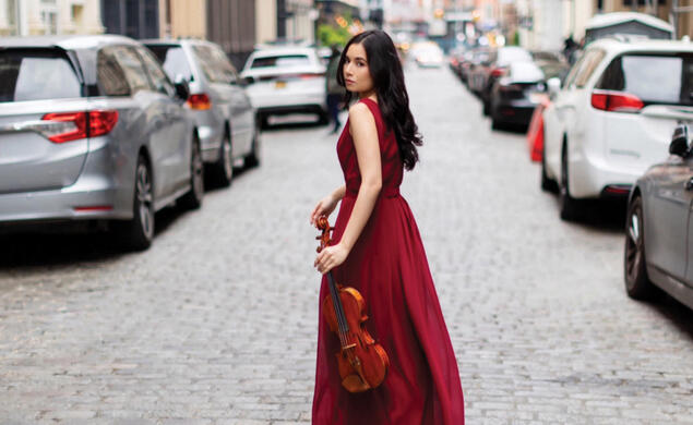 Hana Miyamoto Mundiya ’20 in red dress walking down the middle of the road carrying a violin in New York City
