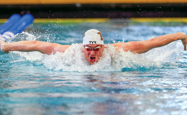 Mitchell Schott competing in a butterfly race in an indoor swimming pool, wearing a swim cap and goggles as water splashes around him during a powerful stroke.