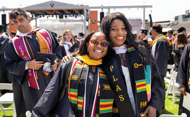 OTC_Commencement Affinity.jpg Two women smile for the camera at a graduation ceremony, wearing multicolored stoles and cords over their black graduation robes.
